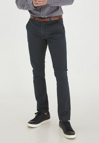 Mørkeblå chinos laget av myk bomull, med slim fit, frontlommer og et brunt lærbelte, parret med svarte sneakers.