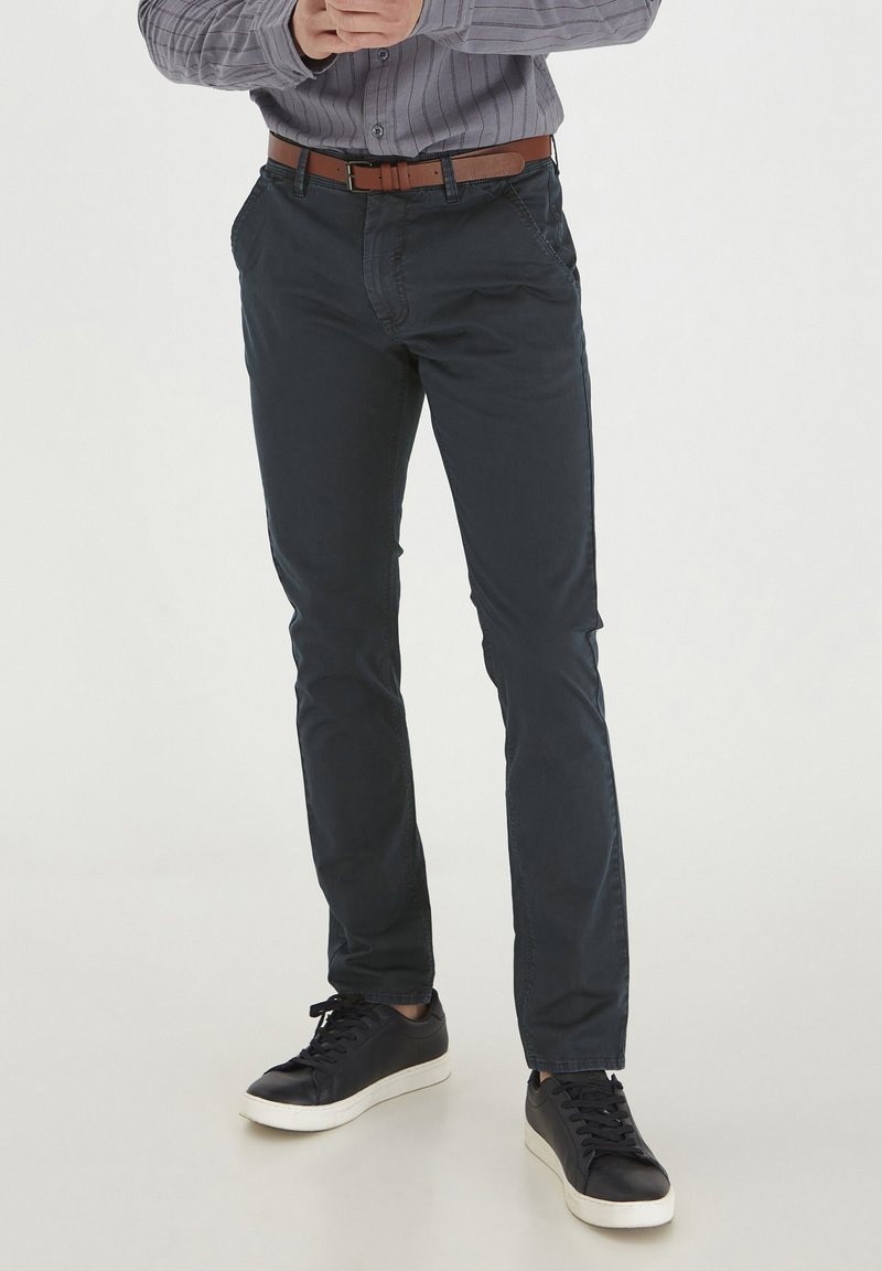 Mørkeblå chinos laget av myk bomull, med slim fit, frontlommer og et brunt lærbelte, parret med svarte sneakers.