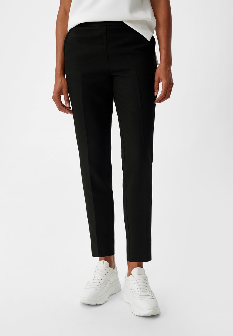 Personne portant un pantalon noir slim et des baskets blanches, debout devant un fond blanc uni.
