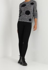 Pull texturé noir et blanc avec de grands pois noirs, associé à des leggings noirs et des bottes noires à lacets. Debout contre un mur blanc.