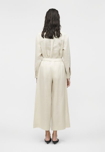 Γυναίκα με σκουρόχρωμα κυματιστά μαλλιά που φοράει κρεμ παντελόνι ψηλόμεσο και φαρδύ, αντίστοιχο πουκάμισο μακρυμάνικο και μαύρα παπούτσια με τακούνι, στραμμένη πλάγια.