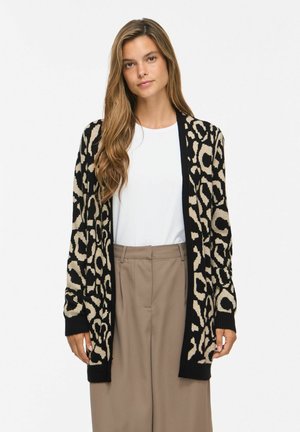 Ung kvinde med langt, bølget hår iført en sort og beige leopardmønstret cardigan over en hvid skjorte og beige bukser, stående foran en ensfarvet baggrund.