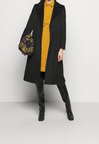 Czarny długi płaszcz z klapami, zestawiony z musztardową sukienką koszulową. Czarne kozaki do kolan oraz torba na sznurki w kratę.