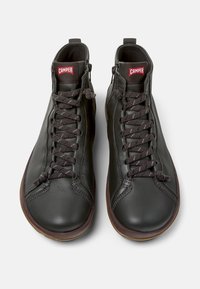 Zapatillas altas de cuero negro con suelas de goma marrón, con un diseño de cordones y un logotipo rojo de "Camper" en la lengüeta.