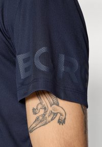 Nära bild av en persons överarm som visar en krokodiltatuering under en marinblå kortärmad t-shirt med delvis synlig text på ärmen.