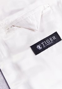 Bílá tkanina s matným leskem obsahuje klopový kapsa. Černá etiketa nese bílý nápis „TIGER OF SWEDEN“ s logem.