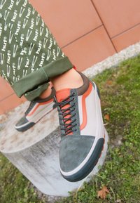 Sneakers med en grön mocka tåhuvud, orange accenter, grå sidopaneler och svarta snören; placerade mot mönstrade gröna byxor och en stenbas.