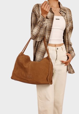 Donna che indossa una camicia a quadri beige, un crop top bianco e pantaloni crema, che tiene una grande borsa a tracolla in pelle marrone con bordi intrecciati.