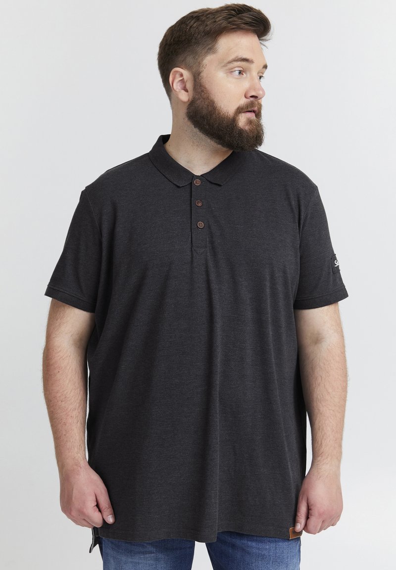 Svart polo shirt med knäppning, korta ärmar och en texturerad yta. Har en logo på ärmen och en liten etikett vid fållen.