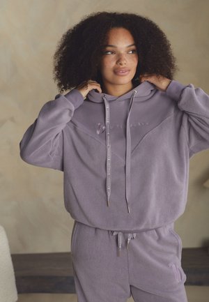 Tânără cu păr creț purtând un hanorac din fleece lavandă și pantaloni de trening asortați, ajustând gluga în interior pe un fundal simplu.