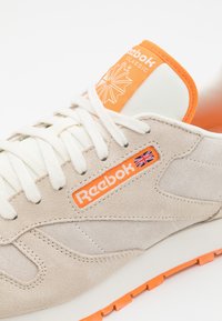 Baskets en daim beige avec une tige texturée, une languette en mesh blanc, des accents orange et un patch logo avec un motif de feuille. Semelle en caoutchouc.