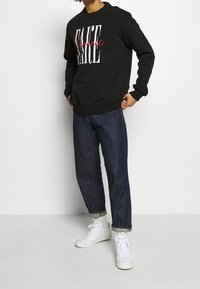 Persona vistiendo un sudadera negra con el texto "PAUSE Romance", jeans oscuros con puños, calcetines blancos y zapatillas blancas frente a un fondo liso.