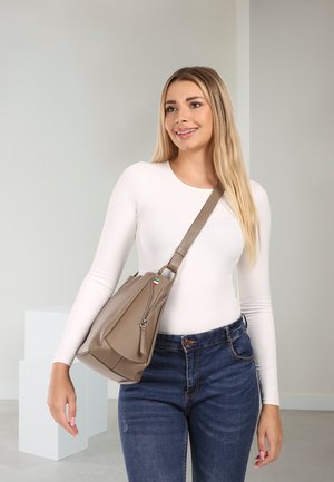 Woman wearing a white long-sleeve top and blue jeans, carrying a taupe leather sling bag over her shoulder, smiling slightly to the side.