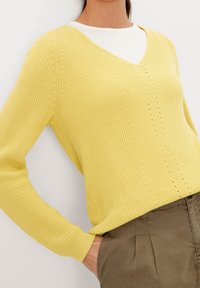 Gelber gestrickter Pullover mit V-Ausschnitt, gerippter Struktur und langen Ärmeln, kombiniert mit einem hellen Unterhemd und olivgrünen Hosen.