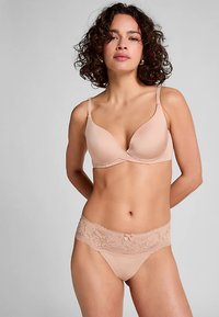 Soutien-gorge rembourré couleur chair avec bretelles réglables et bordure en dentelle, assorti à un string avec détails en dentelle. Les deux pièces présentent une finition lisse.