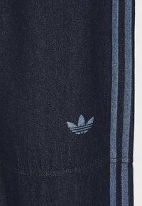 Dark blue denim pants leg with light blue Adidas trefoil logo and two vertical light blue stripes along the side seam.
