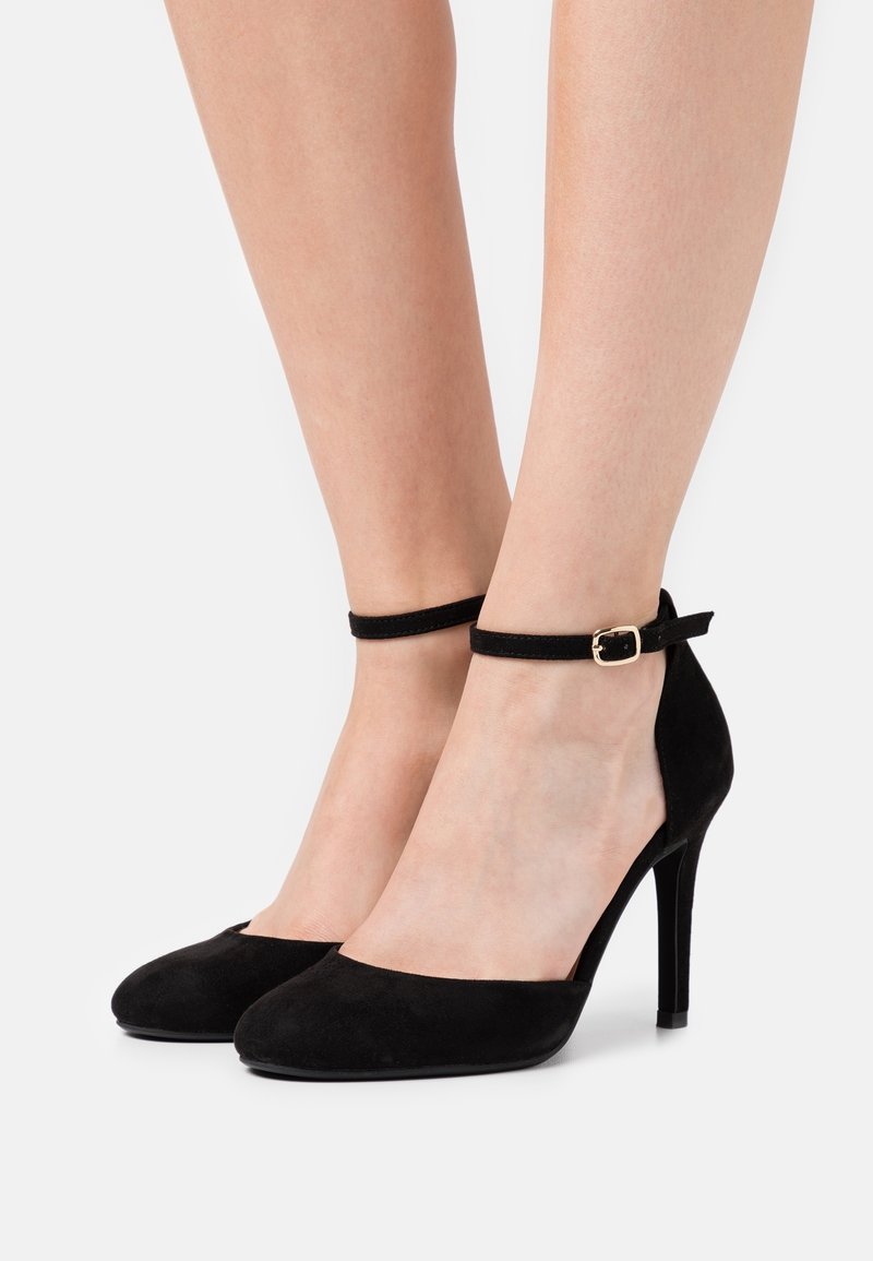 Jambes portant des escarpins en daim noir avec des brides autour de la cheville et des boucles dorées, présentés sur un fond clair uni.