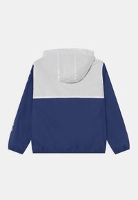 Námořnická modrá větrovka s bílými detaily, kapucí a elastickými manžetami. Má volný střih a lehkou, hladkou texturu.