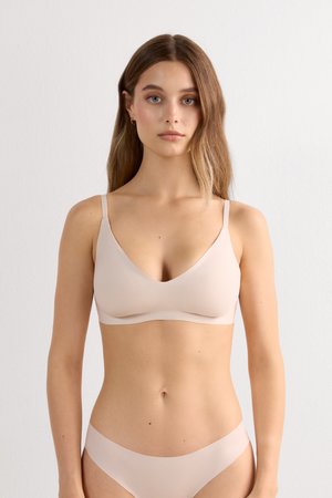 Mujer con el pelo largo y castaño que lleva un sujetador sin costuras color nude y ropa interior a juego, de pie contra una pared blanca lisa, mirando hacia adelante.