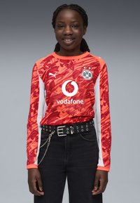 Punainen pitkähihainen jalkapallopaita, jossa on teksturoitu kuvio, valkoiset sivuelementit ja logot. Mustat farkut, joissa on niittivyö ja ketjuyksityiskohta.