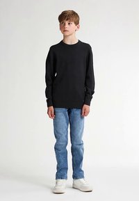 Svart långärmad tröja, jeans och vita sneakers. Ung modell står rak med neutralt ansiktsuttryck mot en enkel ljus bakgrund.