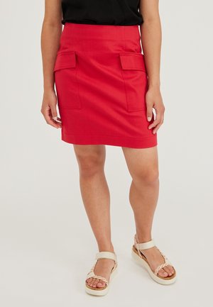 Femme portant une jupe rouge vif avec des poches devant, un haut noir et des sandales ouvertes aux orteils de couleur claire, debout sur un fond uni.