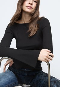 Črna dolgoročna majica z žebljičkastimi dolgimi rokavi in razširjenimi manšetami. Model jo je kombiniral z jeansi, okrašenimi z čipkami, sedi na kromiranem stolu.