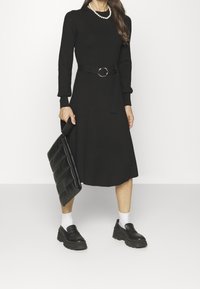 Femme en robe midi noire ceinturée, chaussettes blanches, mocassins à semelle plateforme noirs, collier de perles, tenant une pochette matelassée noire.