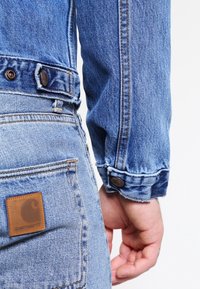 Modrá džínová bunda s manžetami na knoflíky a hnědým koženým záplatou na zadní kapse. Má viditelné švy a volný střih.