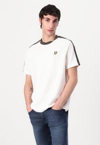 Vit bomull t-shirt med rund halsringning, olivgröna detaljer vid axlarna och en liten gul logotyp på bröstet. Finns i en avslappnad passform.