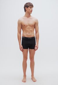 Μαύρα ανδρικά εσώρουχα τύπου μποξεράκια με το λογότυπο Calvin Klein στη λαστιχένια ζώνη μέσης, φορεμένα από έναν άνδρα μοντέλο που στέκεται μπροστά σε απλό φόντο.