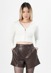 Young woman wearing a white zip-up hooded crop top and high-waisted brown leather shorts with hands in pockets against white background.
