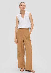 Biała bezrękawnikowa bluzka z dekoltem w kształcie litery V; beżowe spodnie z szerokimi nogawkami z zielonymi haftowanymi wzorami liści z przodu; modelka stoi na neutralnym tle.