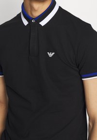 Man i svart polo t-shirt med krage och ärmar i blått, vitt och svart randigt mönster, med en liten vit logotyp på bröstet.