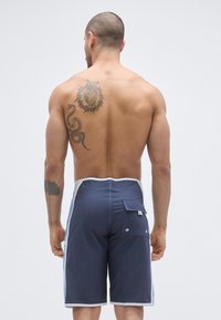 Pantaloni scurți de baie bleumarin cu margine albă, panouri laterale și buzunar în spate. Confecționați din material ușor. Prezintă tatuaje cu șarpe și inimă.