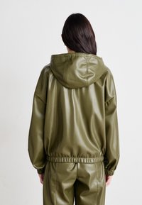 Haut à capuche en simili cuir vert olive avec un cordon de serrage à la capuche, des poignets et une taille élastiques ; texture lisse avec une coupe décontractée.