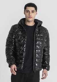 Giovane uomo che indossa un lucido giubbotto imbottito nero con cappuccio sopra una maglietta nera, in piedi contro uno sfondo grigio chiaro uniforme.