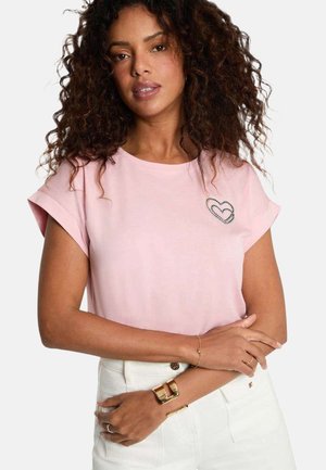 Femme aux cheveux bouclés portant un haut rose clair à manches courtes avec un petit motif double cœur près de l'épaule, un pantalon blanc et des bijoux en or.