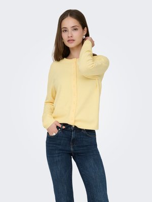 Gele cardigan met een ronde hals en knopen aan de voorkant, gemaakt van zachte stof. Draagt donkerblauwe jeans met een nauwsluitend ontwerp.