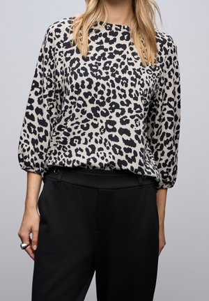 Femme portant un chemisier beige à manches longues avec un imprimé léopard noir, rentré dans un pantalon noir taille haute, sur un fond gris uni.