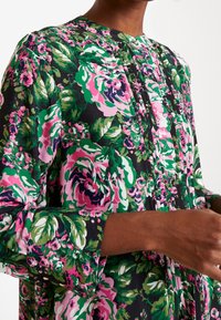 Vestido floral de tejido negro con grandes patrones de rosas en rosa, verde y blanco. Cuenta con un cuello alzado y una tapeta con pliegues verticales.
