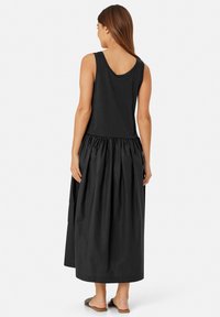 Sort stropløs kjole med tætsiddende top og en fuld, samlet nederdel. Stoffet ser let og glat ud. Flade sandaler synlige.