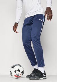 Calças de atletismo em azul-marinho com um corte slim, riscas laterais brancas, acompanhadas de sapatilhas pretas e uma bola de futebol preto e branco.