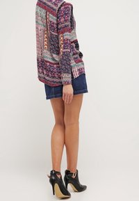 Maglia a maniche lunghe con motivi floreali e geometrici in navy, rosso e verde, abbinata a una gonna di jeans e stivaletti neri alla caviglia.