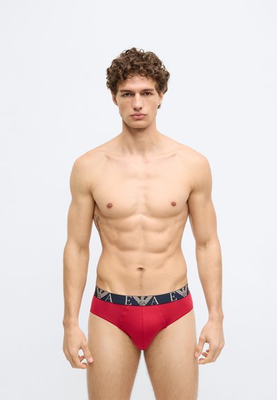 Cueca vermelha de algodão com uma cintura azul-marinho que apresenta um logótipo. Textura suave e design ajustado, realçando a forma do corpo.