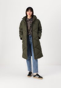 Manteau long matelassé vert olive avec capuche, motif en triangle, fermeture éclair. Porté sur un haut à imprimé léopard, associé à un jean bleu clair et des baskets noires.
