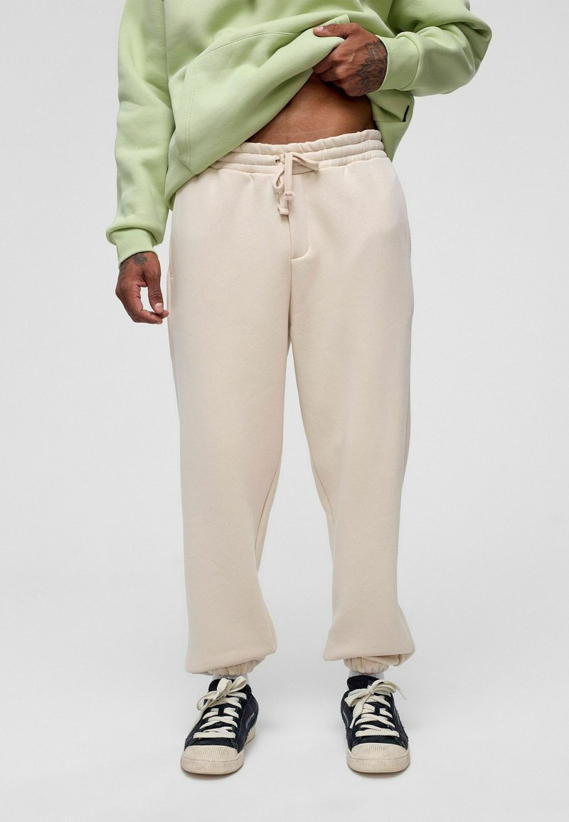 Beige jogger broeken met een elastische tailleband, zijzakken en manchetomslagen. G gedragen met zwart-witte sneakers en een groene hoodie.
