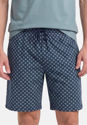 Mann trägt marineblaue Shorts mit weißen kreisförmigen Mustern und einem Kordelzug am Bund, kombiniert mit einem hellblauen Hemd.