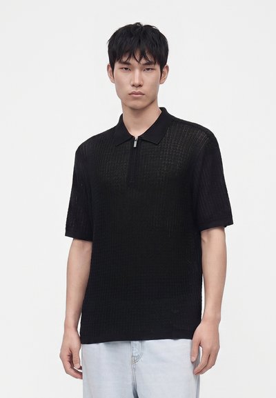 Jeune homme aux cheveux courts et noirs porte un polo en maille noir à manches courtes et un jean bleu clair, debout devant un fond blanc uni.