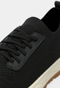 Chaussure de sport noire avec dessus en mesh texturé, lacets plats et semelle en caoutchouc. Présente un patch en cuir lisse sur le côté.